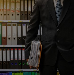 Man holding pile of papers in front of a shelf full of files.